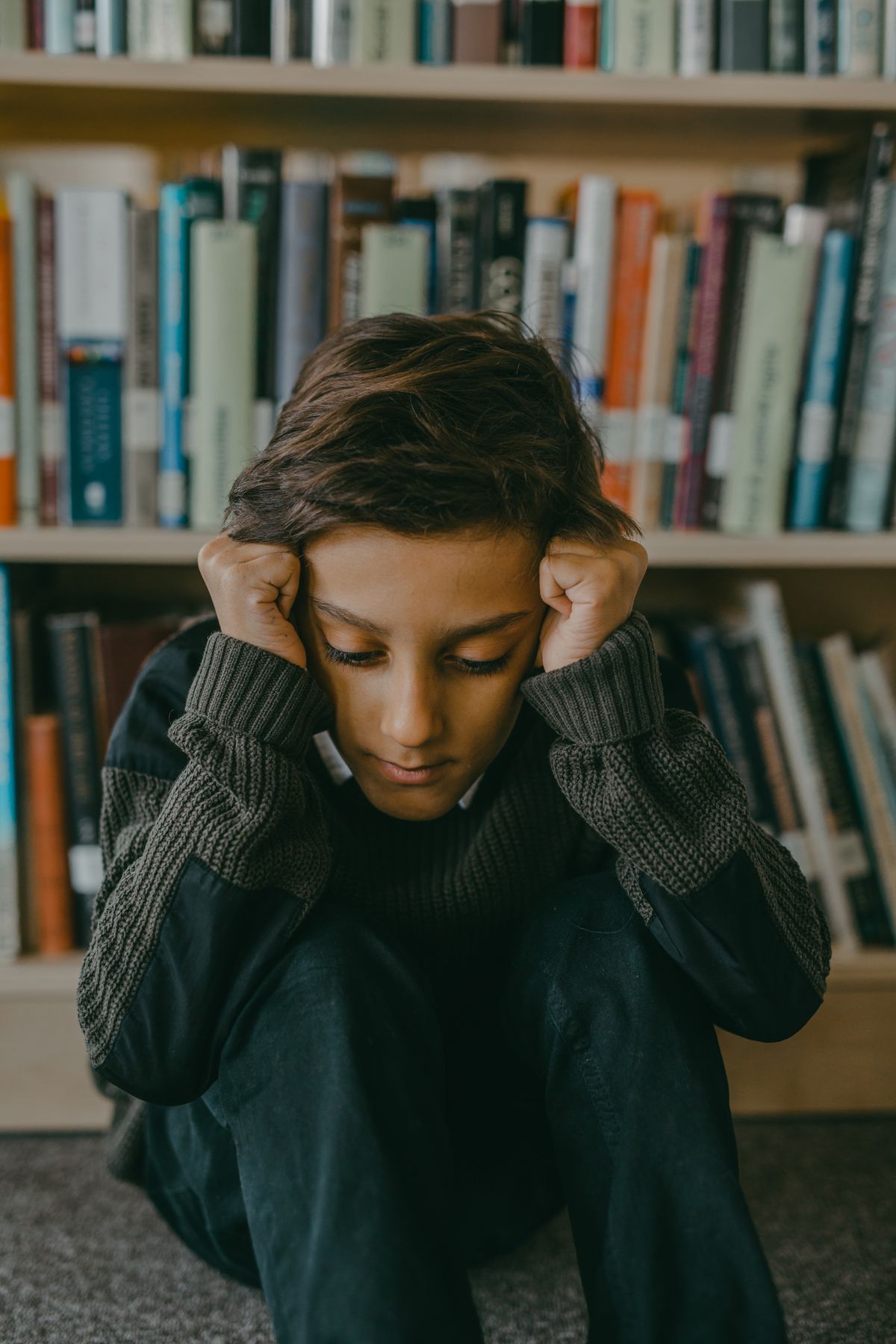 Overwhelmed student sitting against a bookshelf with head in hands — signs of school anxiety in children and teens